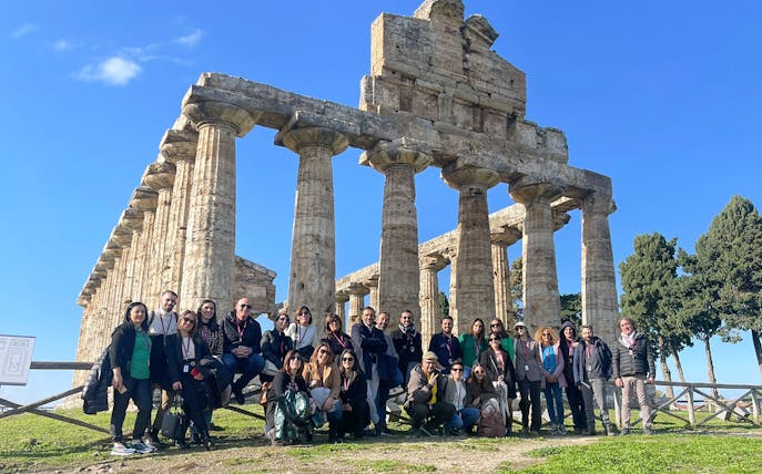 Tour group with guide at Paestum Archaeological Site, Italy, in front of ancient temple ruins.