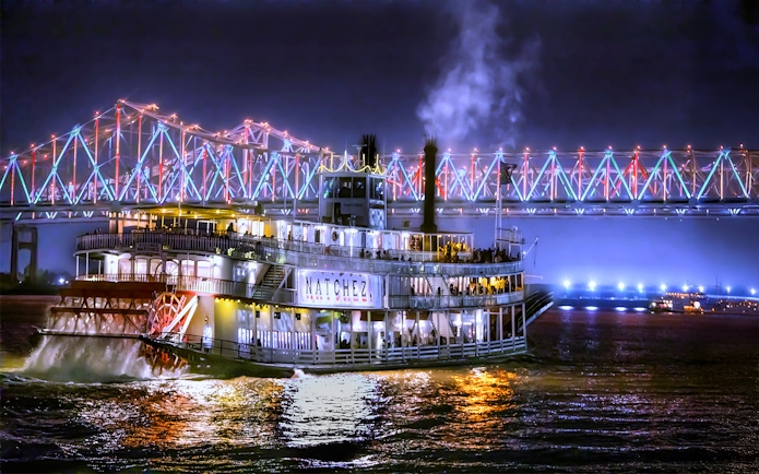 Steamboat Natchez cruising at night under a lit bridge in New Orleans.