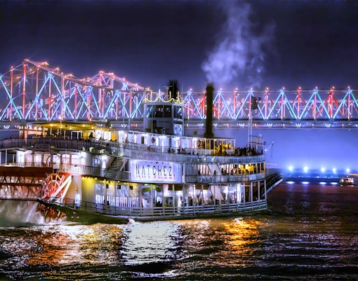 Steamboat Natchez cruising at night under a lit bridge in New Orleans.