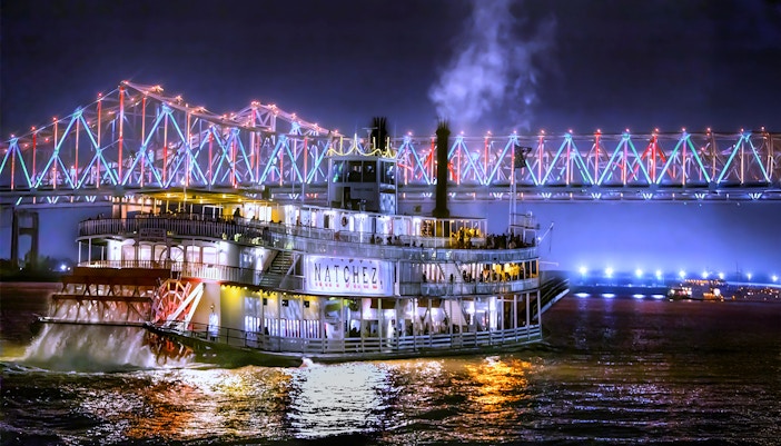 Steamboat Natchez cruising at night under a lit bridge in New Orleans.