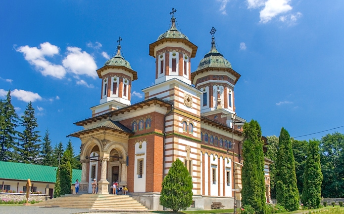 Peles Castle with visitors in front, surrounded by trees, under a clear blue sky.