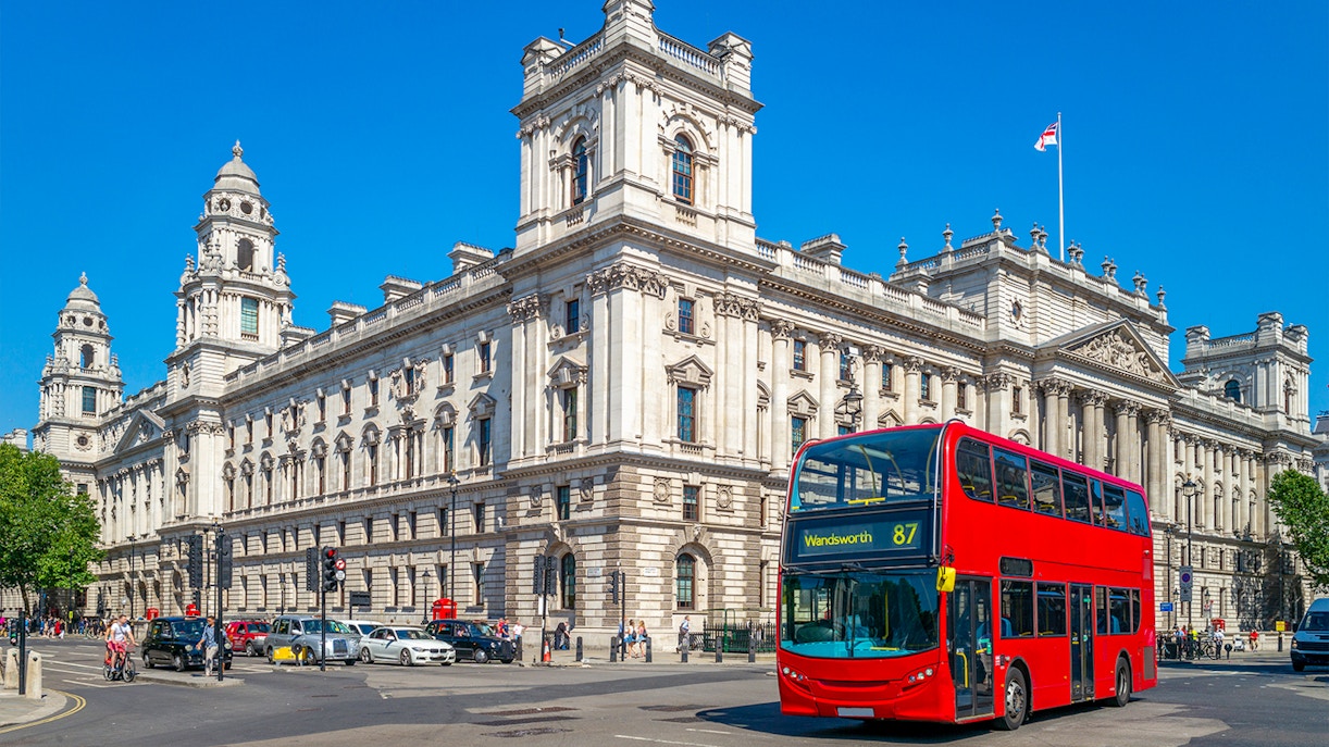 Red double-decker bus on London street near historic building.