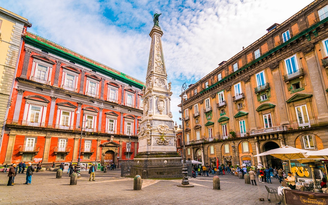 Tourists in Naples exploring historic square with ornate obelisk and colorful buildings.