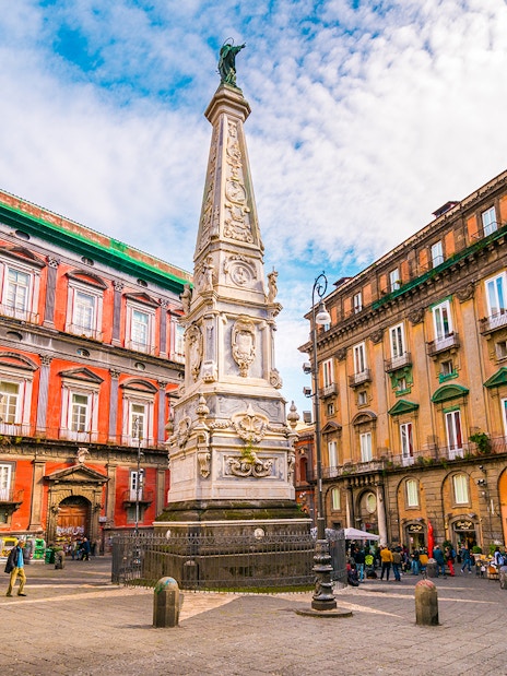 Tourists in Naples exploring historic square with ornate obelisk and colorful buildings.