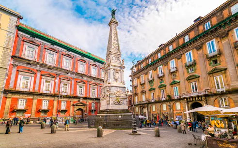 Tourists in Naples exploring historic square with ornate obelisk and colorful buildings.