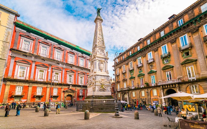 Tourists in Naples exploring historic square with ornate obelisk and colorful buildings.