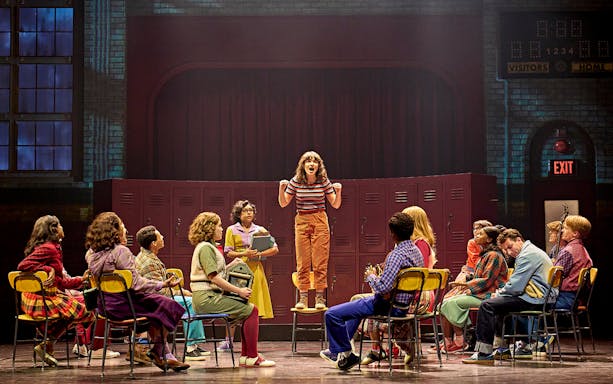 Group of students in a school setting, one standing on a chair, from Stranger Things: The First Shadow.