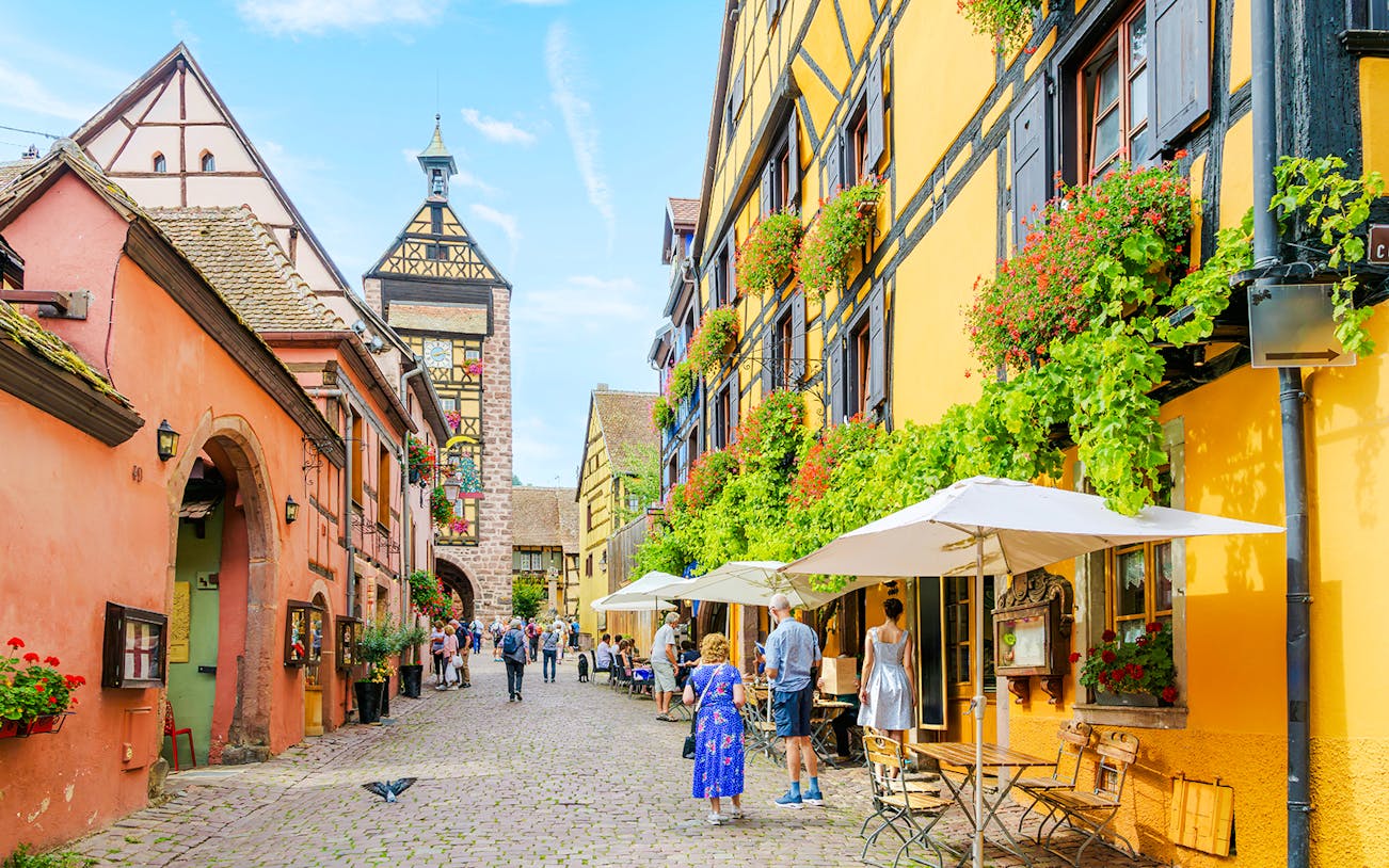 Dolder Belfry Clock Tower in Riquewihr, France, with colorful half-timbered buildings and cobblestone street.