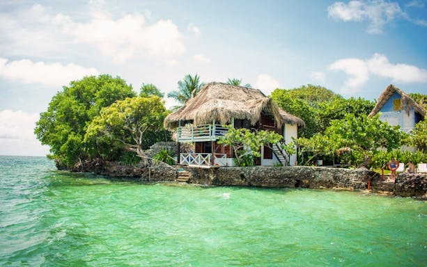 Thatch-roofed hut on Isla del Pirata, Rosario Islands, surrounded by lush greenery and turquoise water.