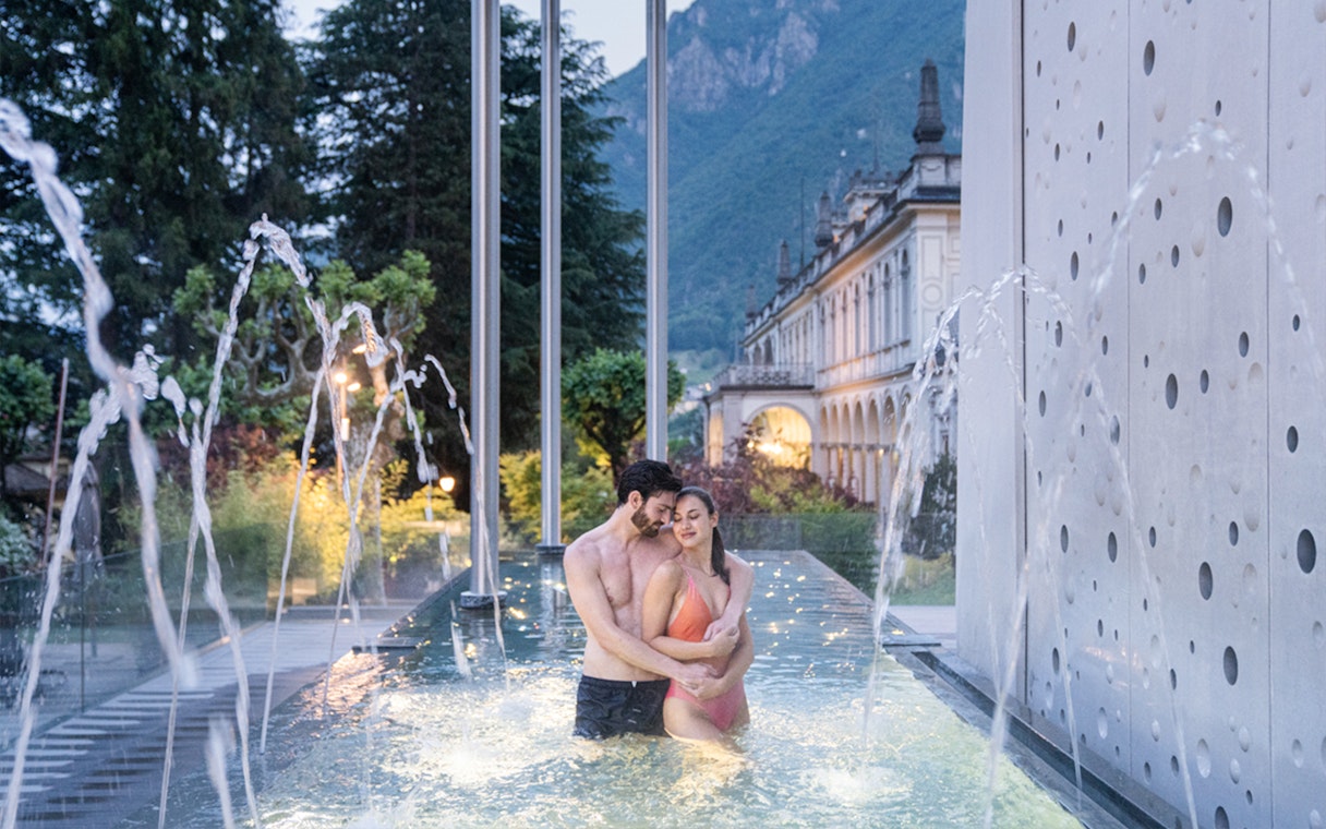 Couple in pool at QC Terme San Pellegrino Resort, Bergamo, with fountains and historic building.