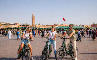 Tourists on e-bikes in Marrakesh's bustling square with Koutoubia Mosque in the background.