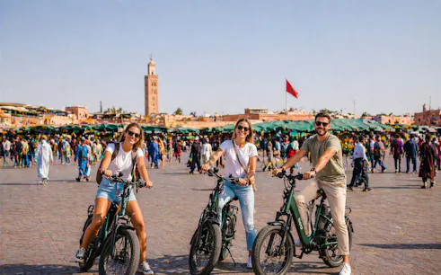 Tourists on e-bikes in Marrakesh's bustling square with Koutoubia Mosque in the background.