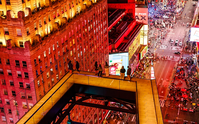 Visitors on a glass walkway overlooking Times Square at night, New York City.