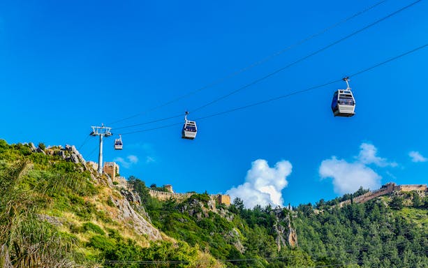 Cable cars ascending to Alanya Castle, surrounded by lush greenery and blue sky.