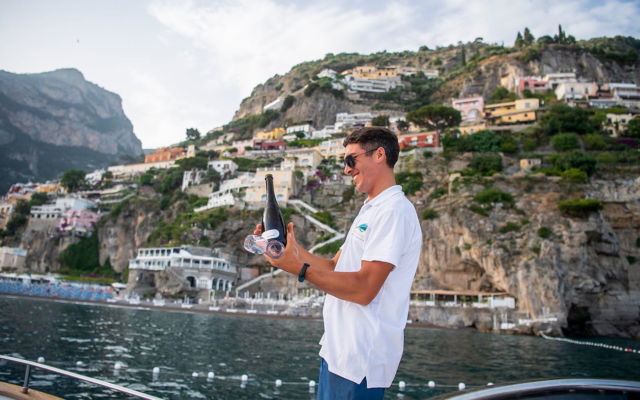 Man holding champagne on a boat with Amalfi Coast cliffs in the background.