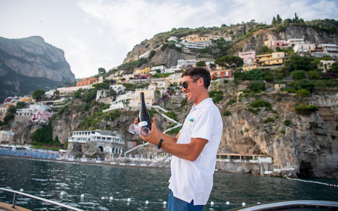Man holding champagne on a boat with Amalfi Coast cliffs in the background.