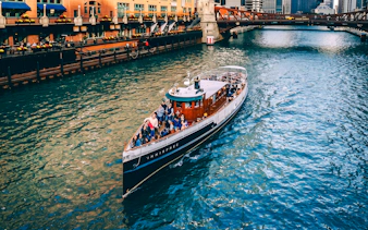 Cruise boat on Chicago River passing under a bridge with city buildings in view.