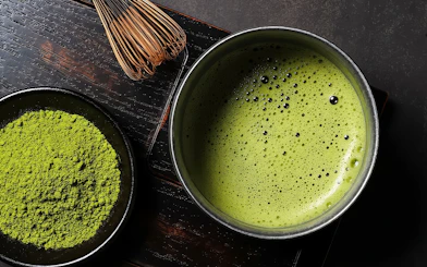 Close-up of matcha powder and whisk next to a bowl of frothy matcha tea.