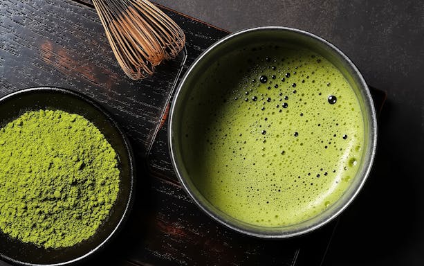 Close-up of matcha powder and whisk next to a bowl of frothy matcha tea.