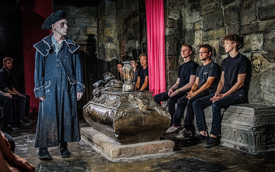 Guide in historical costume leading a tour inside Berlin Dungeon with visitors seated around a tomb.