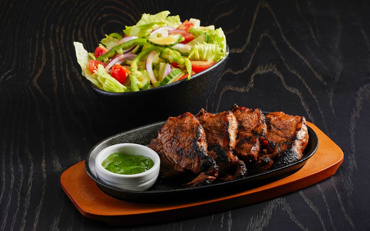 Sliced grilled meat with salad and green sauce on a dark wooden table.
