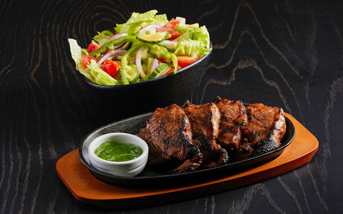 Sliced grilled meat with salad and green sauce on a dark wooden table.