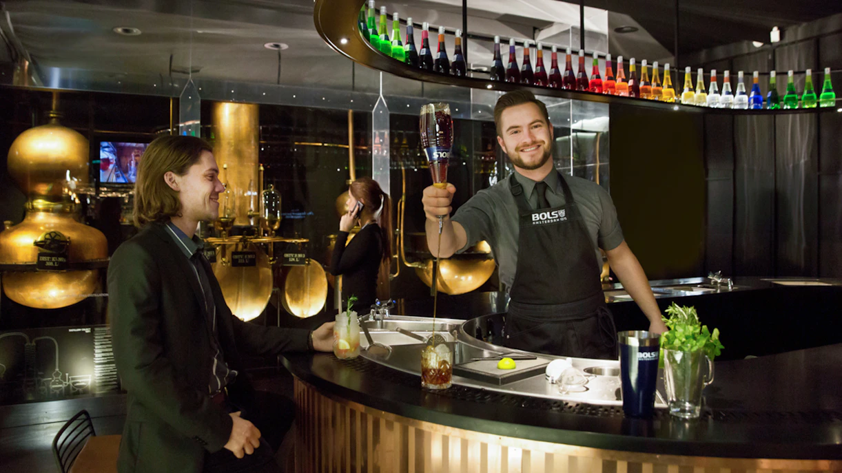 Bartender serving a cocktail at House of Bols in Amsterdam, with colorful bottles displayed above.