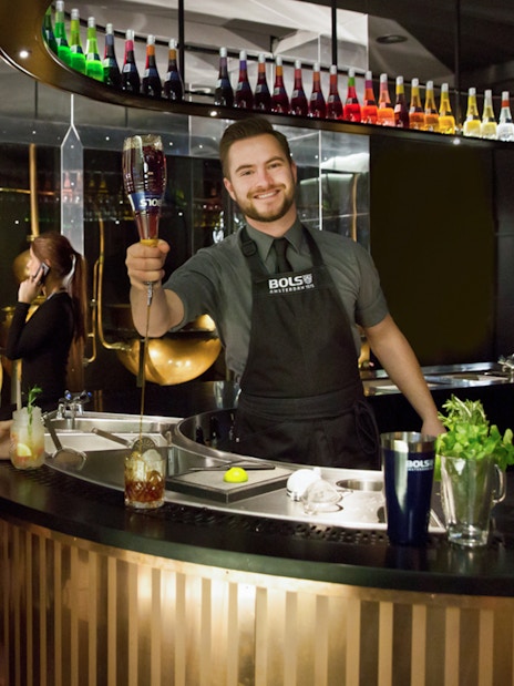 Bartender serving a cocktail at House of Bols in Amsterdam, with colorful bottles displayed above.