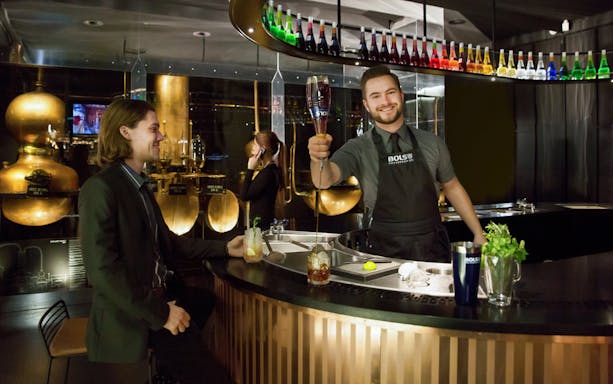 Bartender serving a cocktail at House of Bols in Amsterdam, with colorful bottles displayed above.