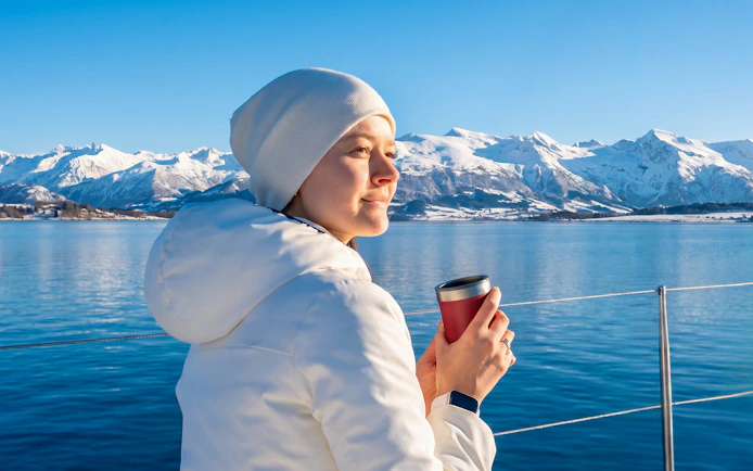 Woman holding a thermo mug on a whale watching cruise in Tromso with snowy mountains in the background.