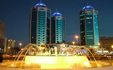 Fountain with illuminated water jets in front of modern skyscrapers in Sharjah at night.