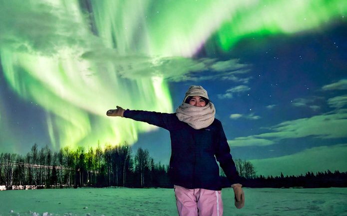 Person pointing at Northern Lights in Kiruna, Sweden, with snowy landscape and trees.