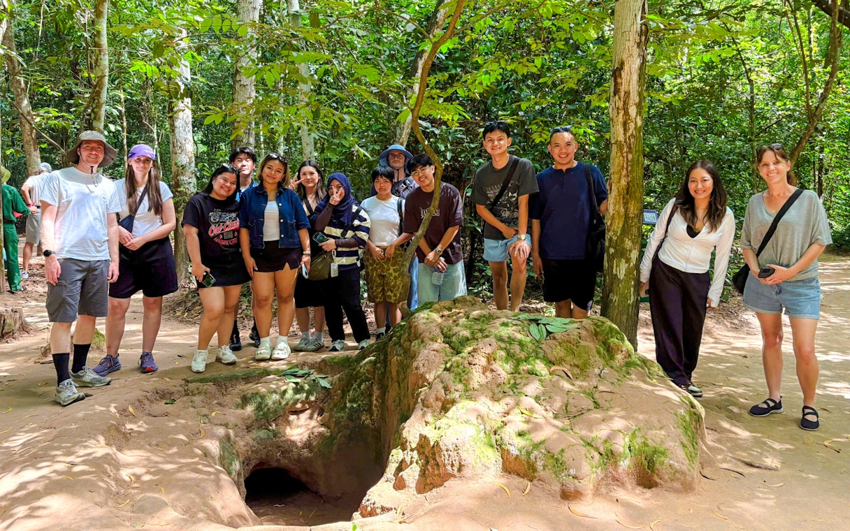 Tourists exploring Cu Chi tunnels with a guide in a forest setting.