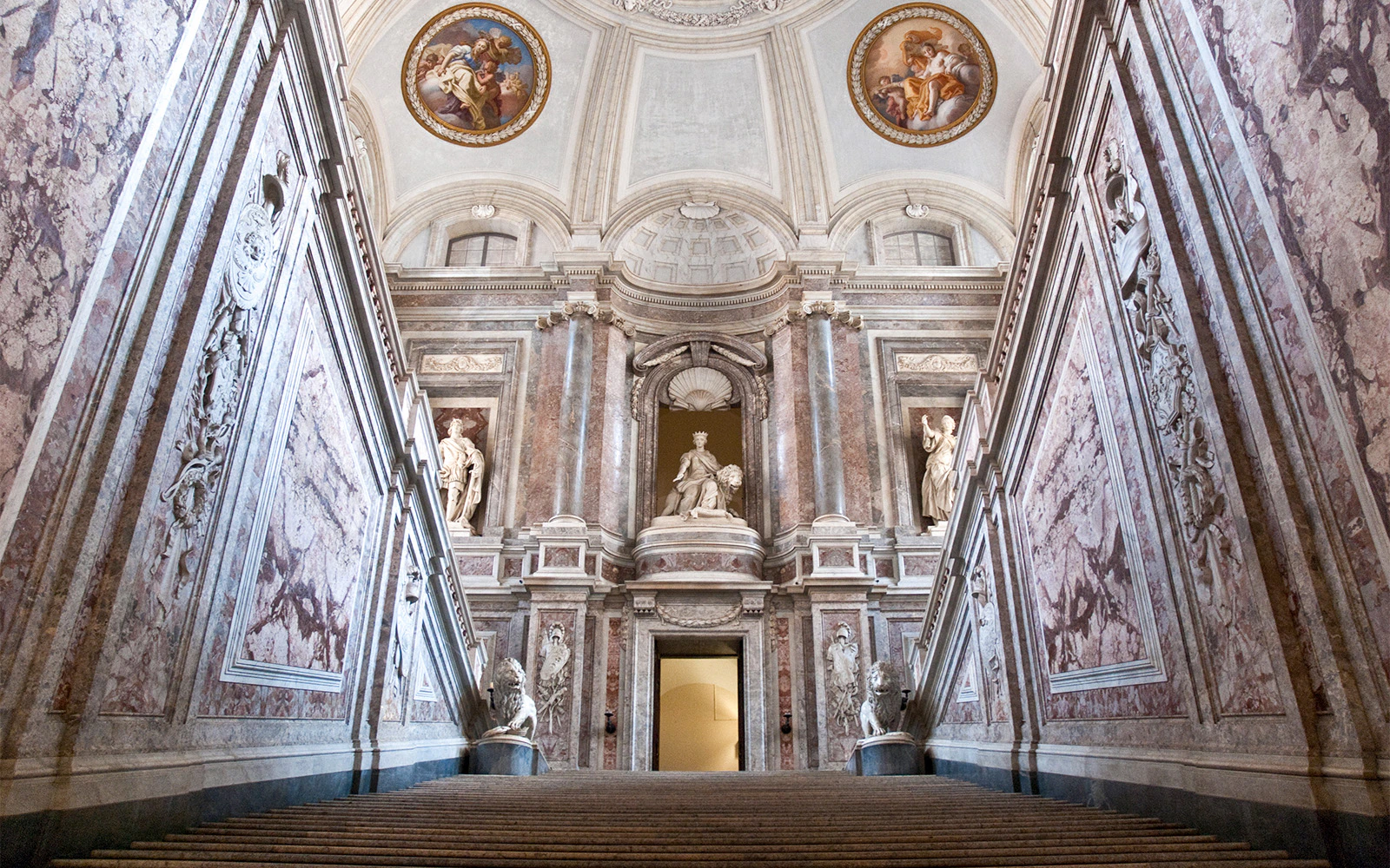 Grand staircase of the Royal Palace of Caserta with ornate marble walls and statues.