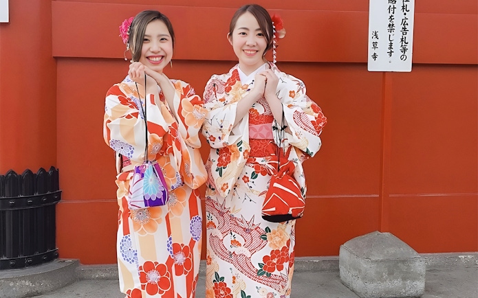 Two people in colorful kimonos smiling in front of a red wall in Asakusa, Tokyo.
