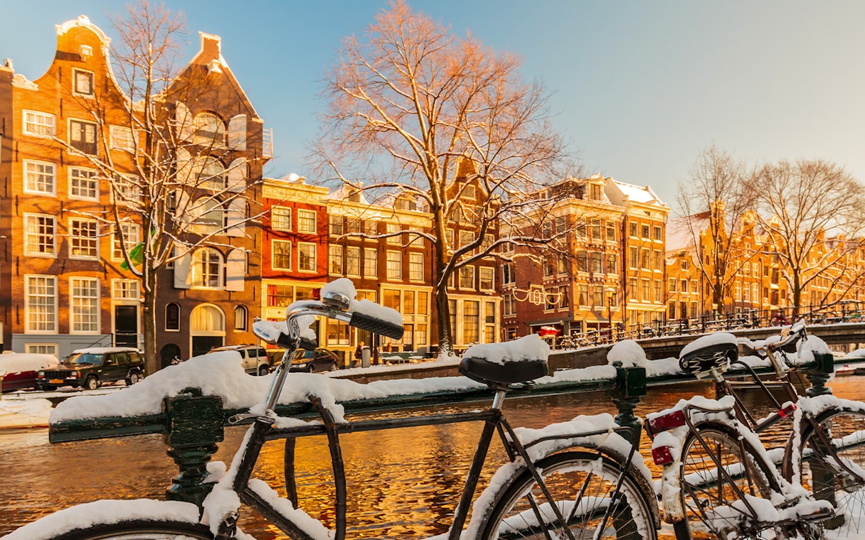 Bicycles covered in snow along an Amsterdam canal with historic buildings in winter.