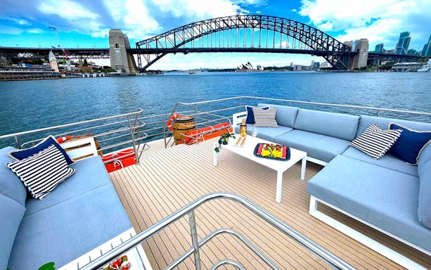 Harbour cruise deck with Sydney Harbour Bridge view, featuring comfortable seating and a fruit platter.