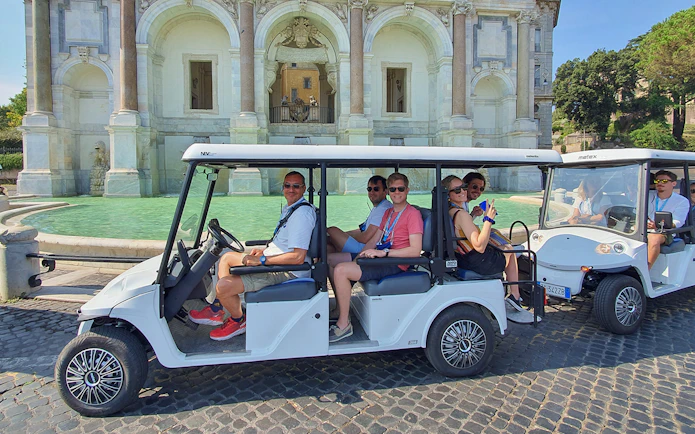 Golf cart with people touring near a historic fountain.