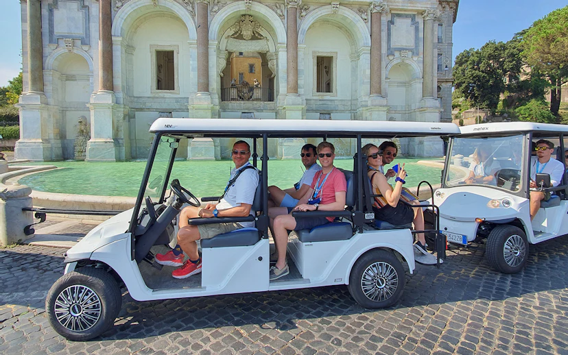 Golf cart with people touring near a historic fountain.