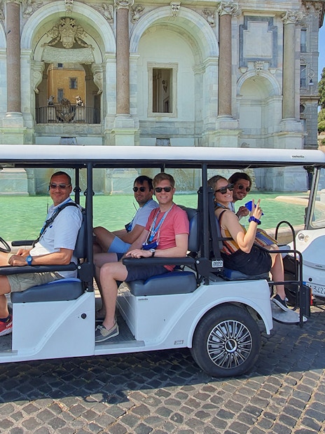 Golf cart with people touring near a historic fountain.