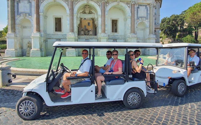 Golf cart with people touring near a historic fountain.