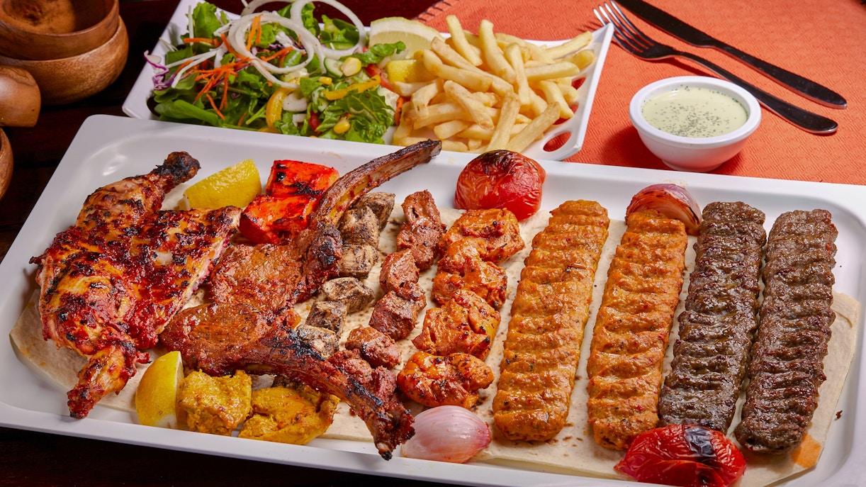 Spicy BBQ mixed grill platter with tikka boti, kababs, fries, salad, and raita on a table.