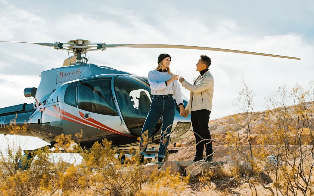 Couple toasting with champagne in front of Maverick Helicopters aircraft in desert setting.