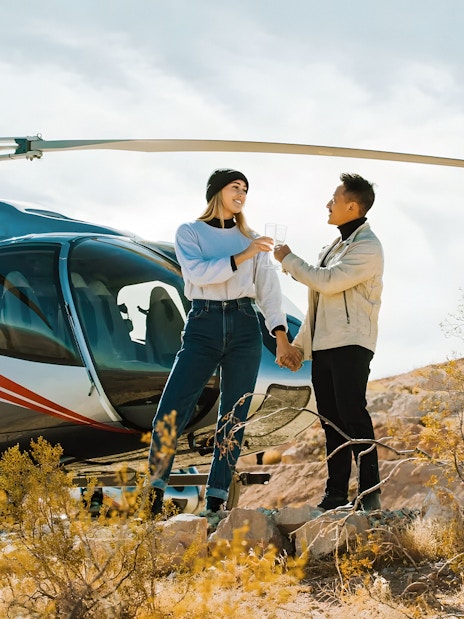 Couple toasting with champagne in front of Maverick Helicopters aircraft in desert setting.
