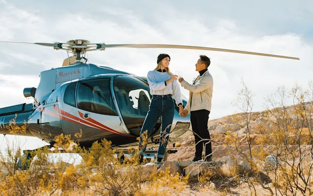 Couple toasting with champagne in front of Maverick Helicopters aircraft in desert setting.