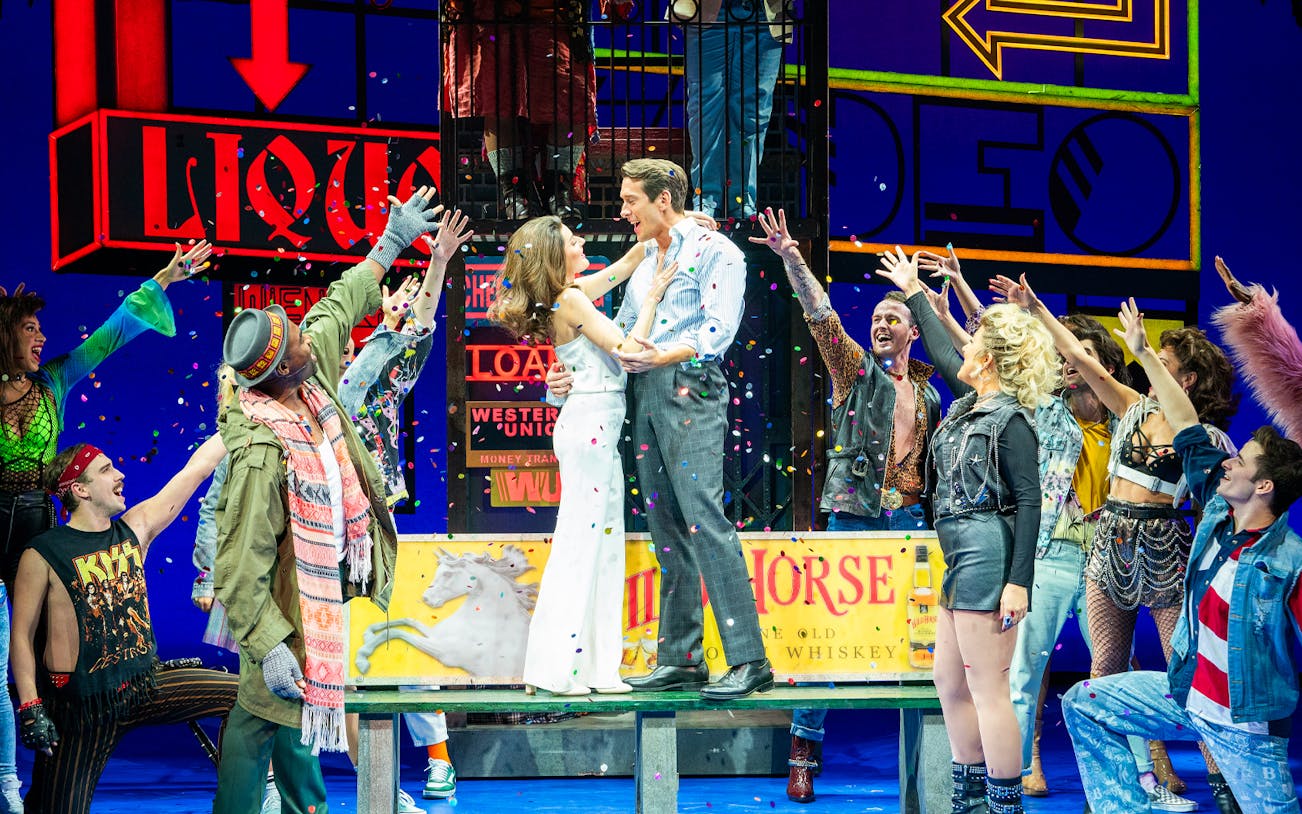 Cast performing a lively scene from Pretty Woman The Musical with colorful neon signs.
