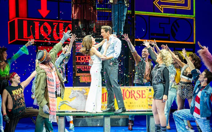 Cast performing a lively scene from Pretty Woman The Musical with colorful neon signs.