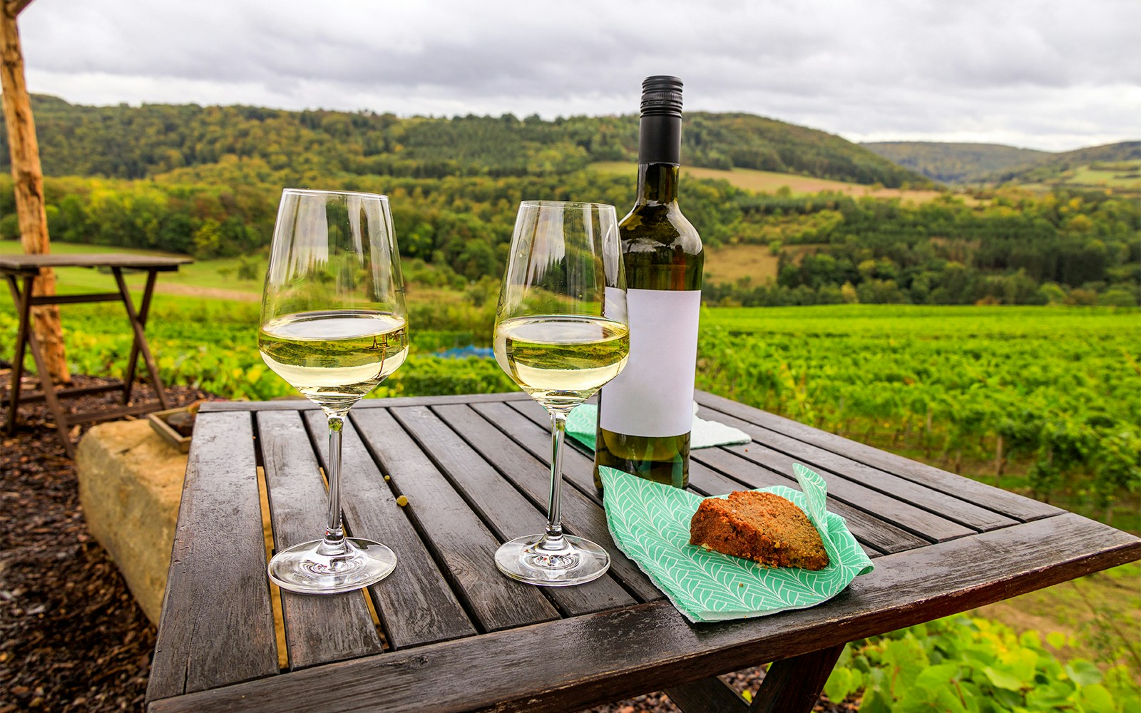 Zwei Gläser Weißwein, eine Weinflasche und ein Stück Kuchen auf einem Klapptisch mit Weinbergen im Hintergrund