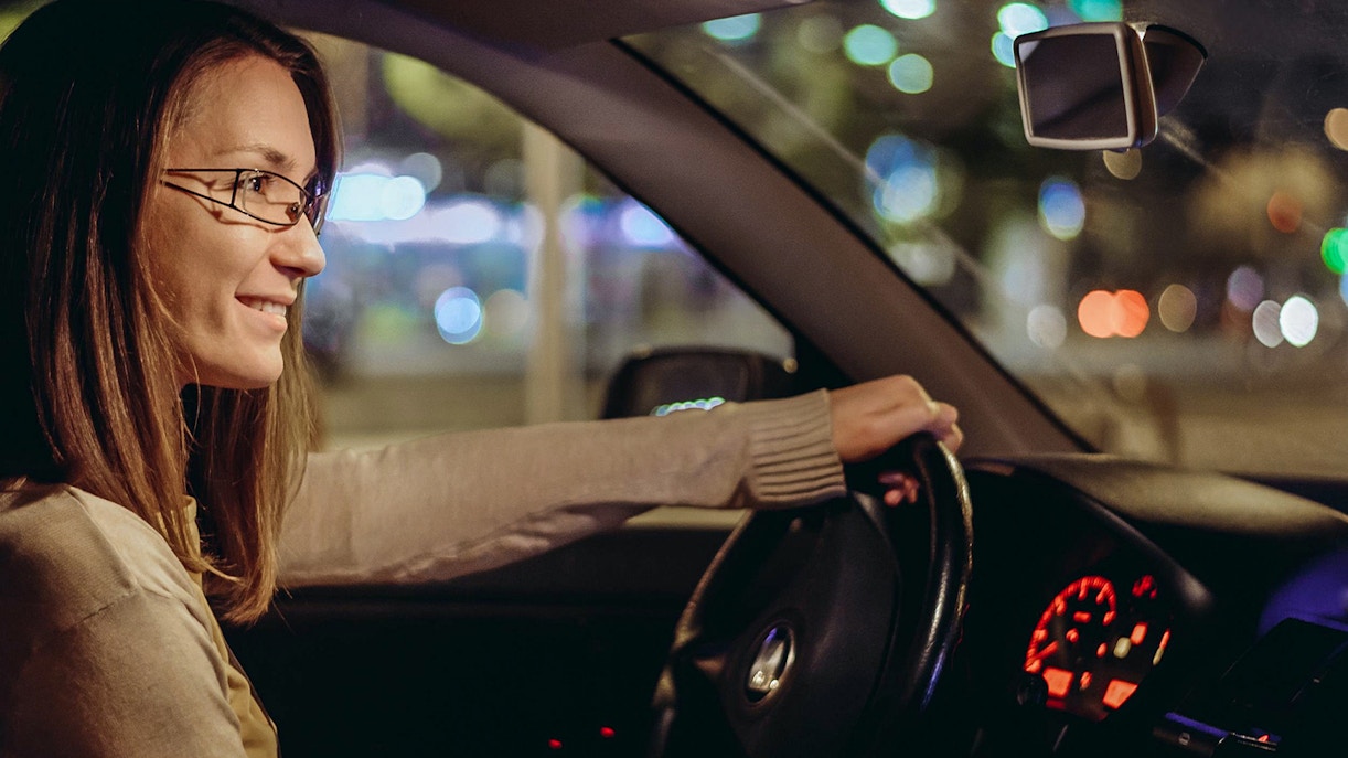 Woman driving a car at night in a city with blurred lights in the background.