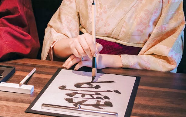 Kimono-clad person practicing Japanese calligraphy during tea ceremony experience.
